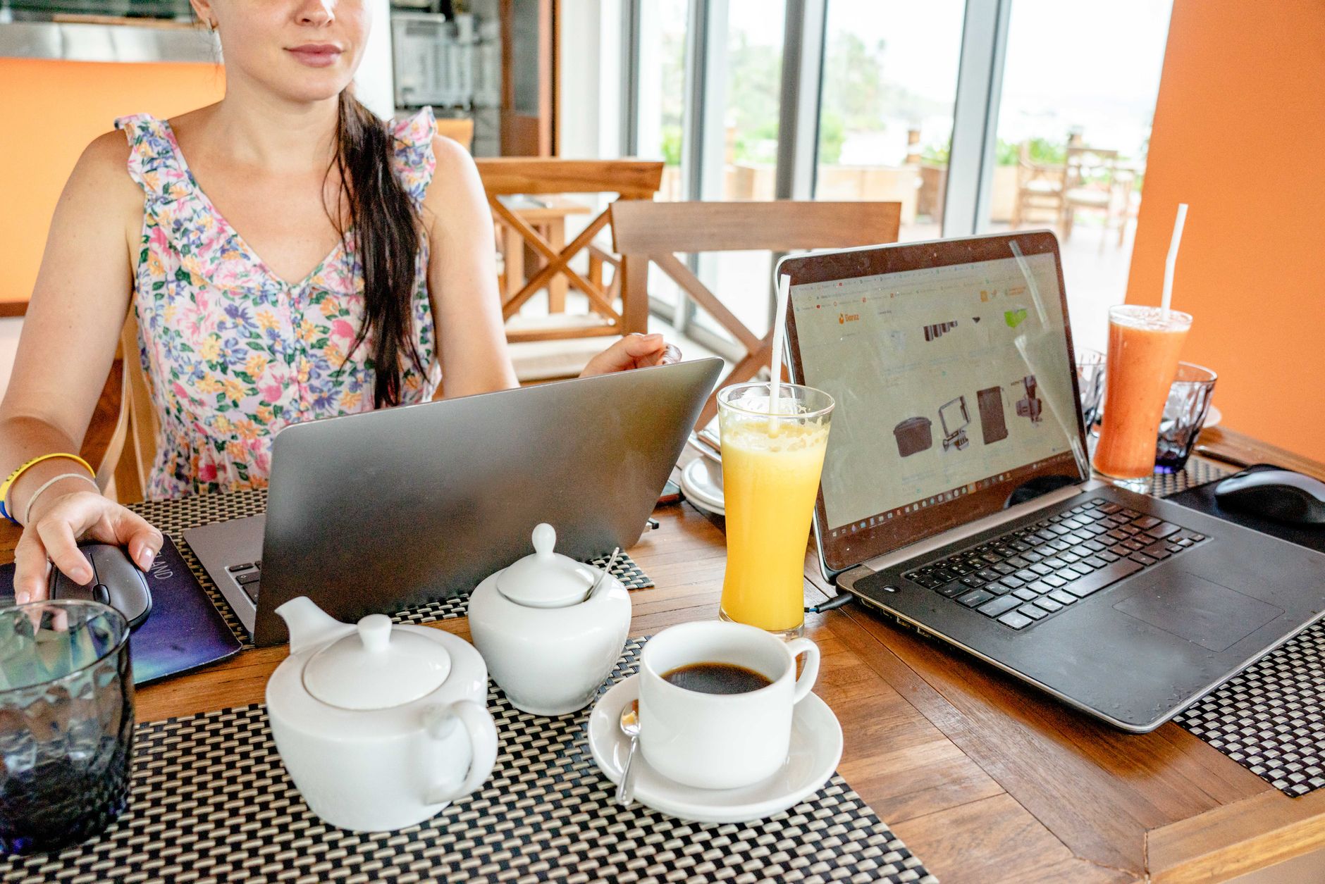 A woman working on laptops at a cafe with drinks and teapots, showcasing a remote work lifestyle.